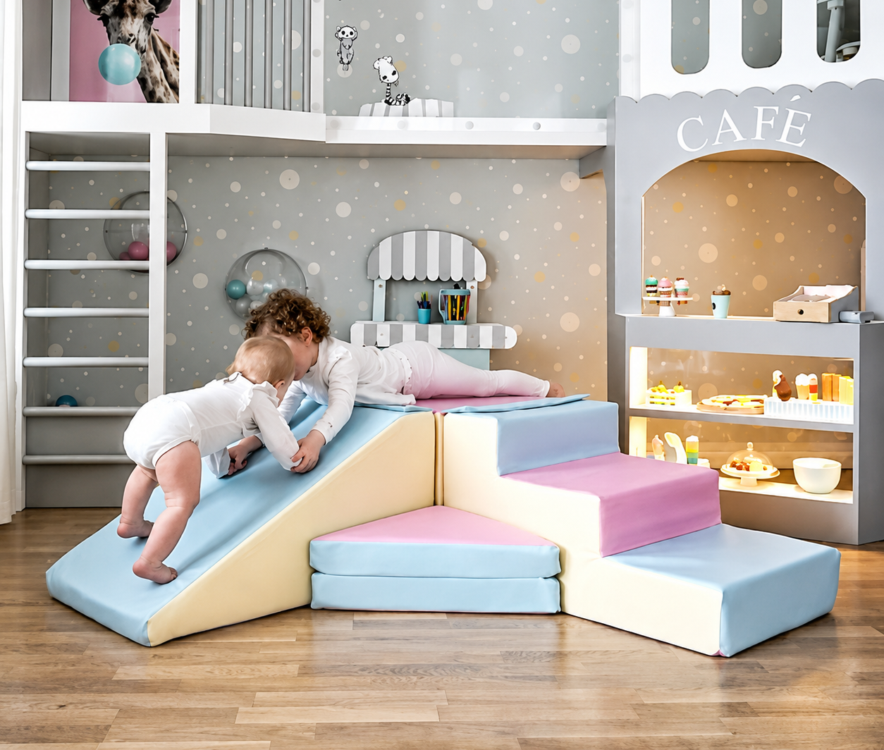 A children's play foam climbing structure in a playroom setting, featuring a light blue and cream slide with two toddlers playing on it, alongside soft foam stairs in light blue and pink.